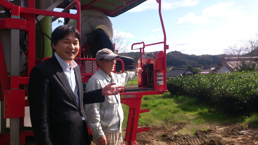 Two men standing next to a red agricultural machine in a matcha field.