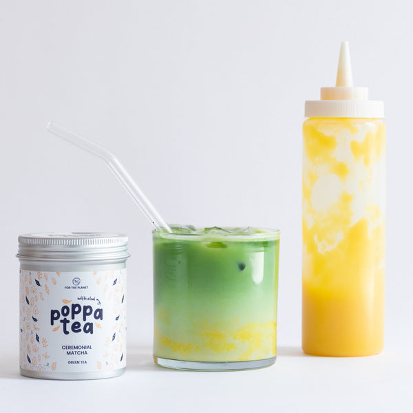 A matcha tea product setup on a clean white background, featuring a white tin labeled 'poppa tea ceremonial matcha with chai' on the left, a clear glass with a green-to-yellow layered iced tea drink with a clear straw in the center, and a tall translucent squeeze bottle filled with yellow drink on the right.