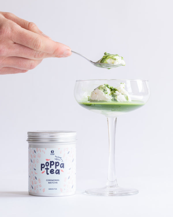 Hand holding a spoon over a glass with a matcha affogato dessert, next to a canister labeled 'Poppa Tea' on a light background