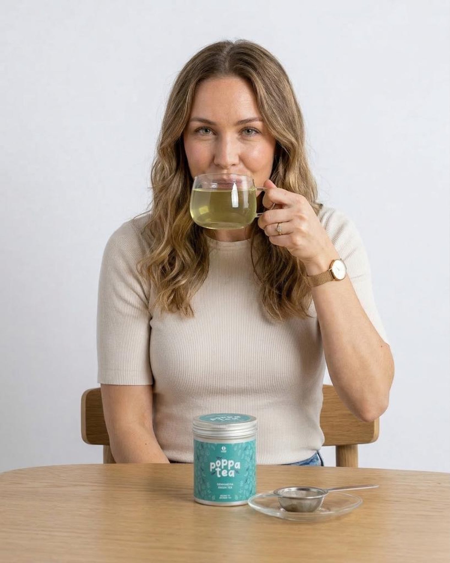 Woman drinking genmaicha tea from a glass with a container of 'Poppa Tea' on a table.