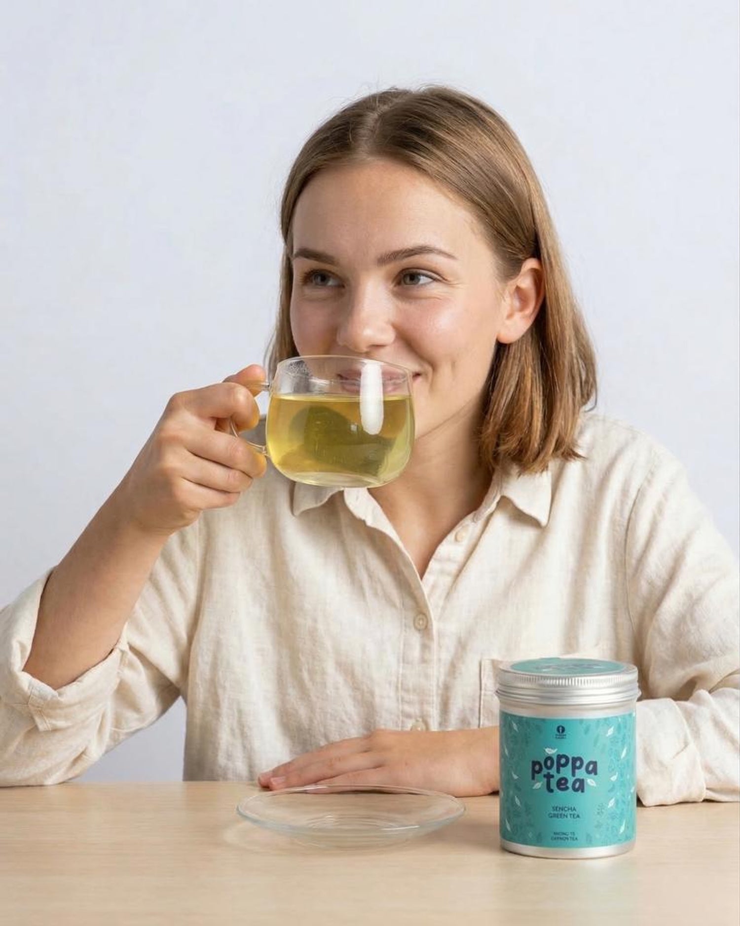 Woman drinking sencha green tea from a cup with a container of Poppa Tea on a table.