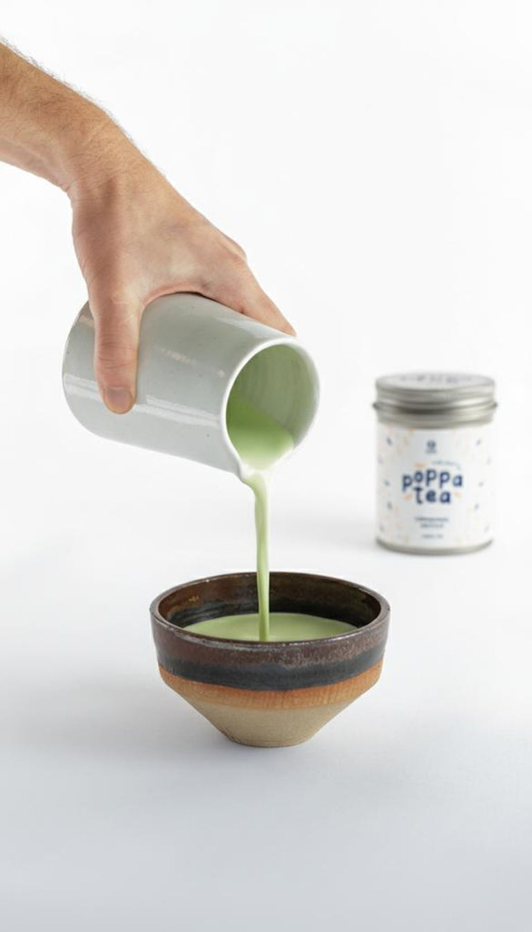 Person pouring green matcha with chai liquid from a small container into a ceramic bowl with a can of 'Poppa Tea' in the background. Making a foamy matcha latte.