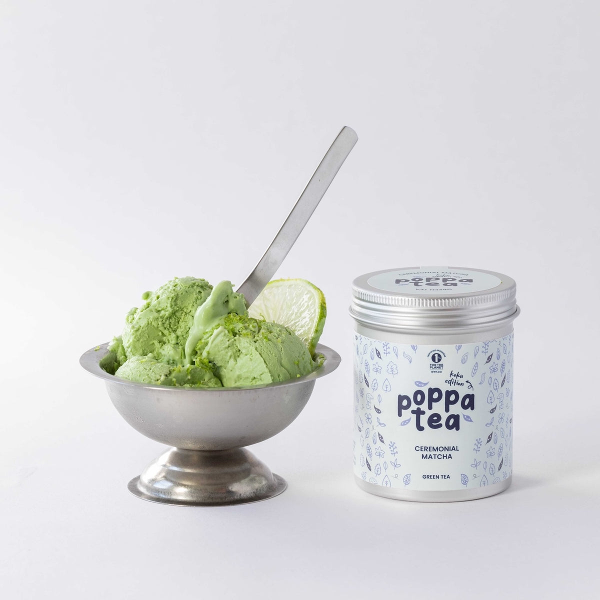 A silver metal tin of PoppaTea Ceremonial Matcha koku edition  stands next to a silver pedestal bowl filled with matcha green ice cream and a lime wedge, on a clean white background.