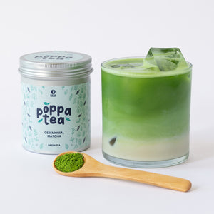 A tin of PoppaTea Matcha green tea sits next to a glass of iced matcha latte with ice cubes, with a wooden spoon holding a scoop of loose matcha in the foreground on a white background.