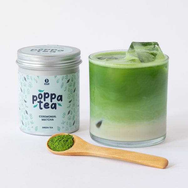 A tin of PoppaTea Matcha green tea sits next to a glass of iced matcha latte with ice cubes, with a wooden spoon holding a scoop of loose matcha in the foreground on a white background.