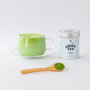 A cup of bright green matcha tea on a clear saucer, a silver-lidded canister labeled 'poppa tea' with 'ceremonial match' on the front, and a wooden spoon holding green matcha powder, arranged on a clean white background.