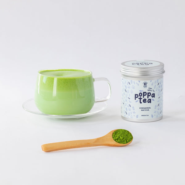 A cup of bright green matcha tea on a clear saucer, a silver-lidded canister labeled 'poppa tea' with 'ceremonial match' on the front, and a wooden spoon holding green matcha powder, arranged on a clean white background.