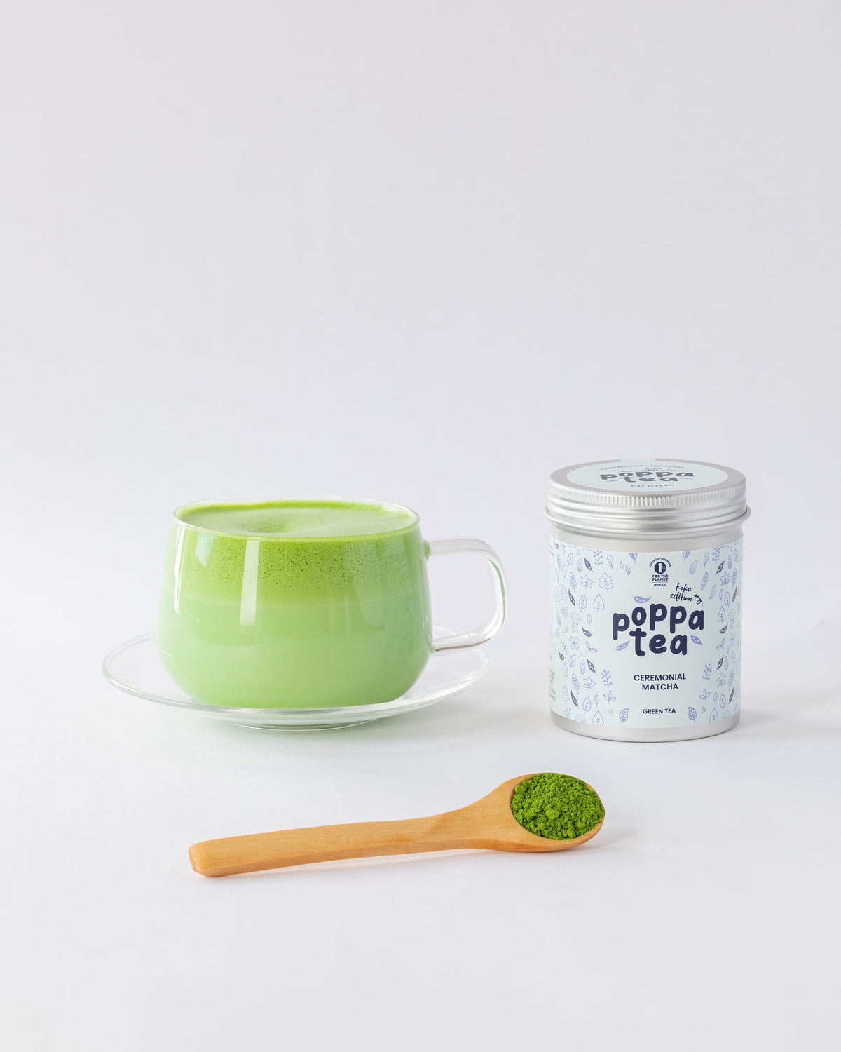 A light green ceramic cup on a saucer with a wooden spoon of green matcha powder, next to a silver canister of Poppa Tea matcha on a clean white background.