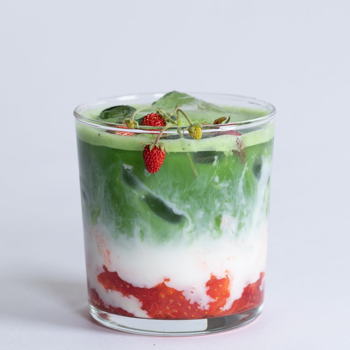 Glass of layered green matcha, white milk, and red drink with berries on a light gray background